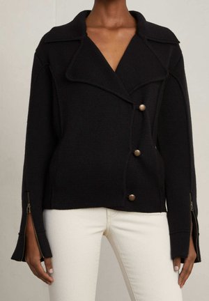 Veste noire texturée à boutons avec large col et fermetures éclair sur les manches longues, portée avec un pantalon blanc.