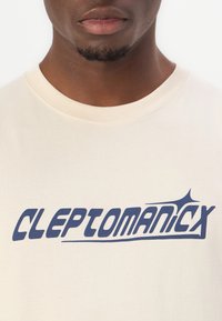 Cream-colored cotton t-shirt featuring bold blue text "CLEPTOMANICX" with a stylized accent on the letter "X."