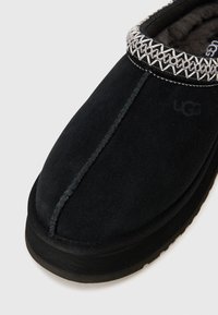 Juodos suede šlepetės su pilkšvu siuvinėjimu, siūlėmis ir patvariu juodu guminio padu. Matomas žymios prekės ženklo logotipas.