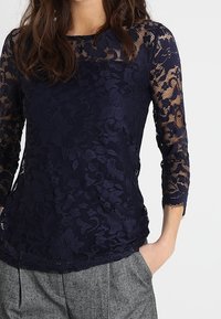 Námořnická modrá krajková blůza s tříčtvrtečními rukávy, zdobená květinovými vzory a průhlednou texturou, kombinována se šedými kalhotami.