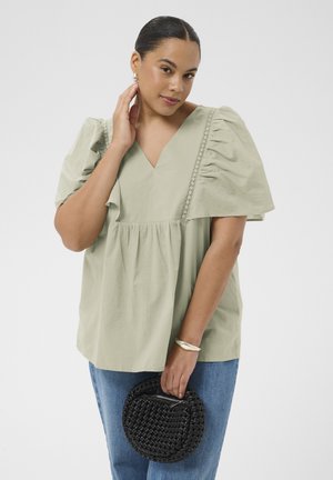 Kobieta z zaczesanymi do tyłu włosami, ubrana w jasnozieloną bluzkę z bufkami i niebieskie dżinsy, trzymająca czarną plecioną, okrągłą torebkę.