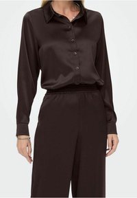 Femme portant une chemise noire en satin à manches longues, boutonnée et rentrée dans un pantalon noir en satin taille haute, accessoirisée avec un bracelet et un collier.