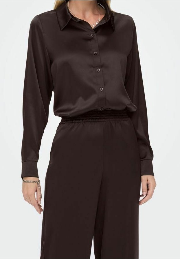 Femme portant une chemise noire en satin à manches longues, boutonnée et rentrée dans un pantalon noir en satin taille haute, accessoirisée avec un bracelet et un collier.