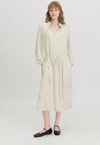 Helles beige Langarmkleid mit V-Ausschnitt und verstellbarem Taillenbund, mit Seitentaschen und gerafften Ärmelbündchen, getragen mit schwarzen flachen Schuhen.