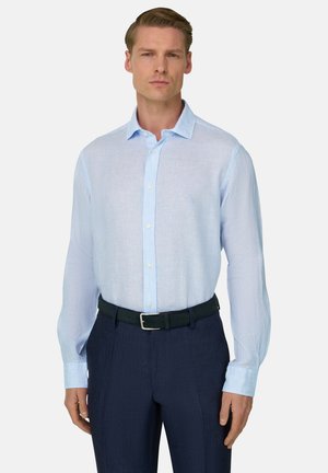 Mężczyzna ubrany w jasnoniebieską, pasiastą koszulę z długim rękawem, wsuniętą w ciemnogranatowe spodnie z czarnym paskiem, stojący na jednolitym tle.
