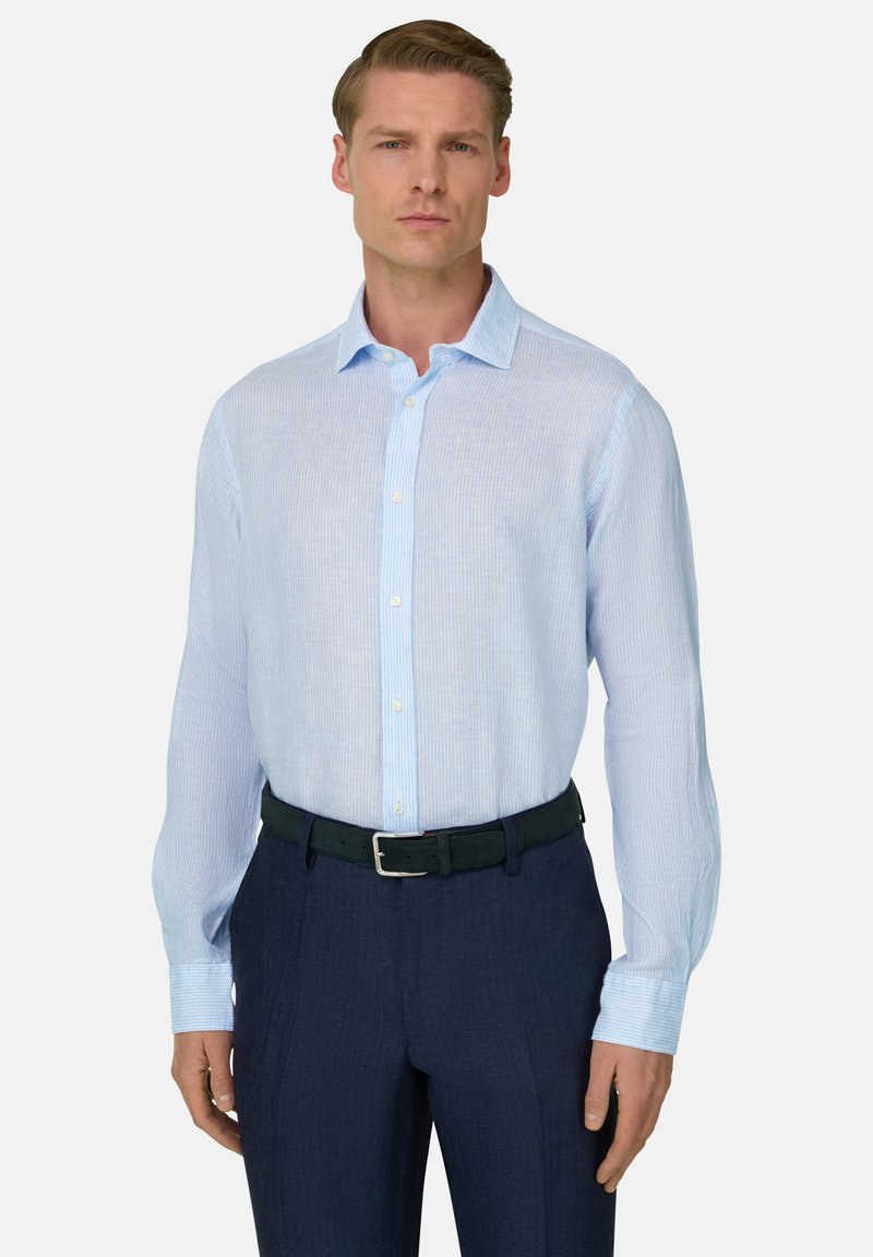 Homme portant une chemise à rayures bleu clair à manches longues, rentrée dans un pantalon bleu marine foncé avec une ceinture noire, debout devant un fond uni.