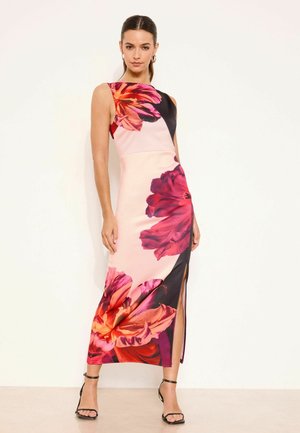 Kobieta w bezrękawowej, dopasowanej maxi sukience z dużymi czerwonymi i różowymi kwiatowymi wzorami oraz rozcięciem sięgającym uda, w połączeniu z czarnymi szpilkami.