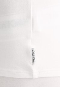 Tricou alb din bumbac, cu decolteu rotund și mâneci scurte, având un mic etichetă cu logo-ul Calvin Klein pe cusătura laterală.