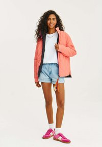 Růžová prošívaná bunda s černým interiérem, nošena přes bílé tričko a džínové šortky, doplněná růžovými teniskami a barevnými třásněmi na šortkách.