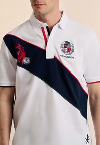 Camisa polo blanca con un panel diagonal azul marino, acentos rojos, logotipos bordados y costuras en contraste en el cuello y las mangas.