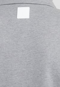 Tela texturizada gris claro con una etiqueta cuadrada blanca cosida cerca del centro superior en la parte trasera de una prenda.