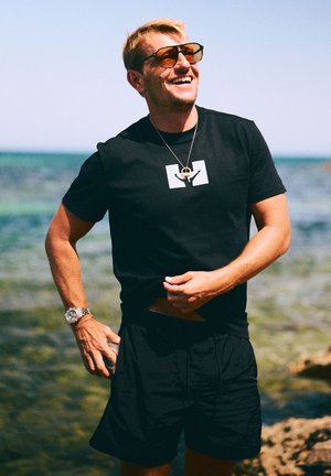 Schwarzes T-Shirt mit weißem Logo, schwarze Shorts, silberne Uhr und goldene Halskette an einem Mann im Freien, der vor einer Strandkulisse lächelt.