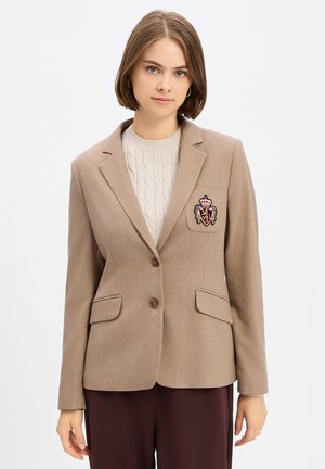 Jonge vrouw met kort bruin haar die een beige blazer draagt met een wapenembleem en daaronder een crème kabelgebreide trui.