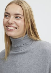 Grijze coltrui met ribstructuur en aansluitende kraag, gedragen door een glimlachende persoon met lang, steil blond haar.