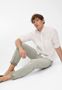 Camicia leggera di lino bianco con tasca, abbinata a pantaloni grigi chiari arrotolati. Modello seduto, indossa scarpe beige slip-on.