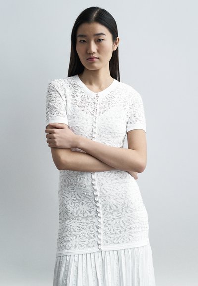 Jeune femme aux longs cheveux foncés portant un haut blanc en dentelle à manches courtes avec des boutons, debout les bras croisés devant un fond gris.