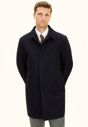 Homme portant un pardessus noir, une chemise blanche et une cravate marron, debout les mains derrière le dos sur un fond clair uni.