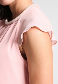 Blouse rose avec un tissu doux et léger, des manches évasées et un détail de col en perles. La texture semble lisse et fluide.