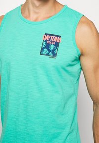 Top de tirantes color turquesa que presenta un gráfico colorido de "Daytona Beach" con palmeras; textura suave, diseño sin mangas y escote redondo.