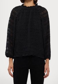 Blusa nera a maniche lunghe con tessuto strutturato e arricciato. Collo alto con un dettaglio arricciato e vestibilità ampia. Abbinata a pantaloni neri.