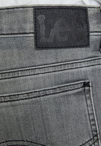 Jean en denim gris avec une étiquette en cuir noir texturé portant le logo "Leo", poches cousues et détails de couture visibles.