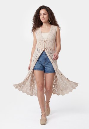 Femme marchant portant un long gilet beige en crochet sur un haut sans manches, un short en jean et des chaussures compensées beiges avec des brides à la cheville.