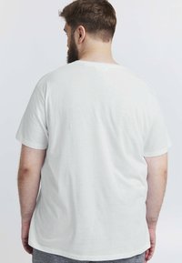 Mand med kort brunt hår og skæg iført en simpel hvid t-shirt med korte ærmer, som vender væk mod en ensartet lys baggrund.