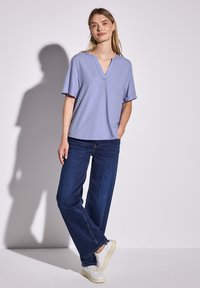 Top viola chiaro a V con maniche corte, abbinato a jeans scuri a gamba larga e sneakers bianche. Tessuto morbido, vestibilità rilassata, stile casual.