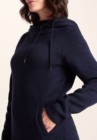 Navy blue pullover with a soft texture, featuring a cowl neck, drawstrings, and side pockets. Fitted sleeves and modern design.
