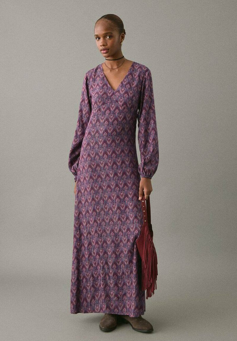 Rochie maxi din material purpuriu cu tipar, având guler în V, mâneci lungi și un design texturat. Modelul ține o geantă cu franjuri color burgund.