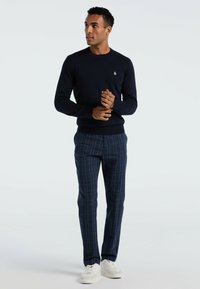 Marineblauwe gebreide trui met een ronde hals, gecombineerd met marineblauwe broeken met een lichtblauw ruitpatroon. Witte sneakers.