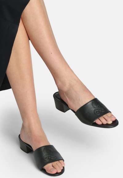 Jambes croisées d'une femme portant des mules à talons ouvertes en cuir noir avec un motif embossé sur un fond blanc.