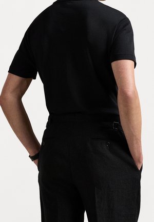 Person, die ein schwarzes kurzärmeliges Hemd und schwarze Hose trägt, mit den Händen in den Taschen, von hinten vor einem einfarbigen Hintergrund gezeigt.