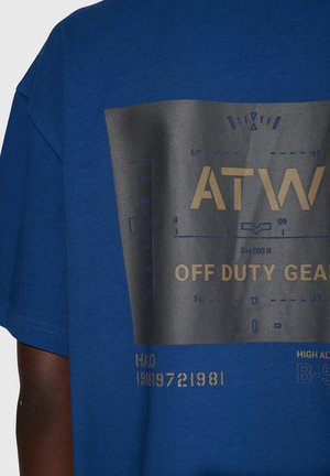 Niebieska bawełniana koszulka z dużym czarnym nadrukiem graficznym oraz złotym tekstem i wzorami, w tym "ATW" i "OFF DUTY GEAR." Krótkie rękawy.