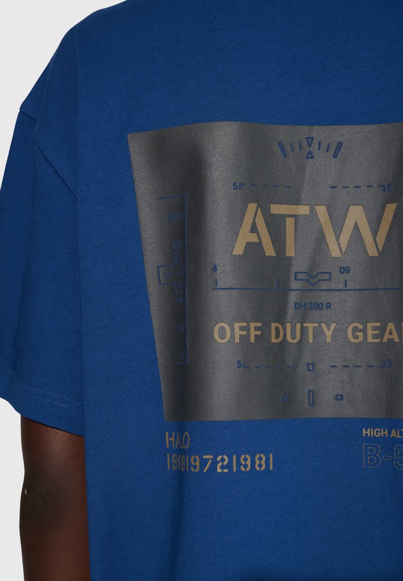 Blå bomuld t-shirt med et stort sort grafisk print med gyldne tekster og motiver, inklusive "ATW" og "OFF DUTY GEAR." Kortærmet.