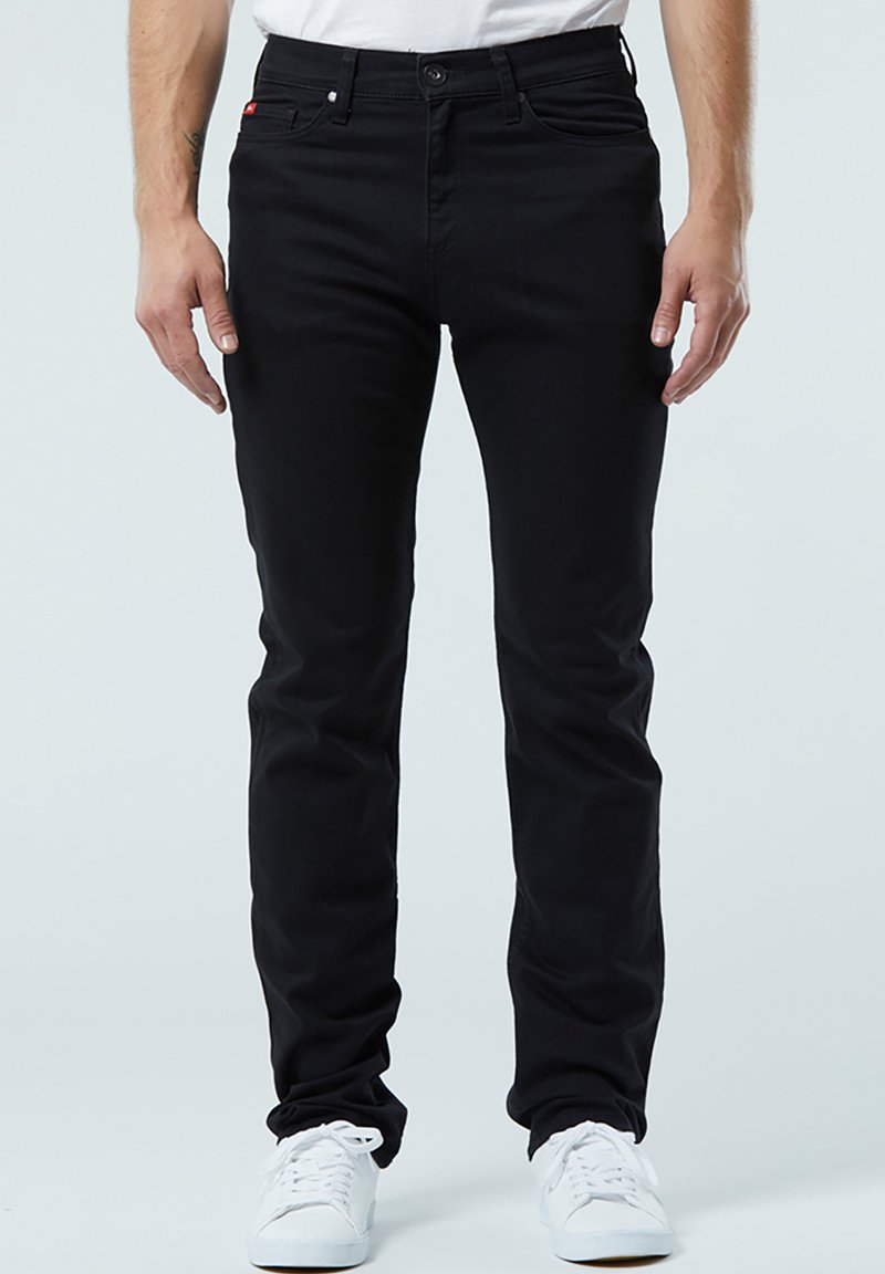 Schwarze Denim-Hosen im Slim-Fit, mit fünf Taschen und Metallknopfverschluss. Getragen mit einem weißen T-Shirt und weißen Sneakern.