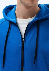 Sudadera azul con cremallera, hecha de un tejido suave, con una cremallera y cordones negros. Incluye una camiseta blanca debajo con un estampado gráfico.