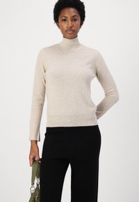 Svetar s turtleneck iz svetlo bež pletene tkanine, z dolgimi rokavi in rebrastimi manšetami in robom. V kombinaciji z črnimi hlačami.