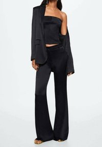 Donna che indossa un top nero a spalla unica abbinato a pantaloni a gamba larga a vita alta e un blazer nero coordinato appoggiato su una spalla.