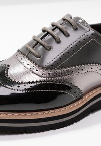 Chaussure à bouts fleuris noire et argentée avec une finition brillante, accents métalliques texturés, lacets gris et semelle superposée contrastante.