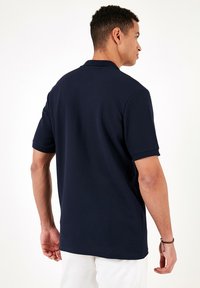 Cămașă polo bleumarin cu țesătură texturată, cu mâneci scurte și guler clasic, prezentată din spate cu o linie de hem dreaptă.