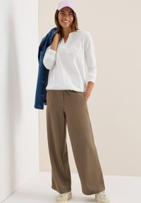 Femme souriante portant une casquette rose, un haut à manches longues blanc, un pantalon large marron, des baskets, tenant une veste en jean sur son épaule.
