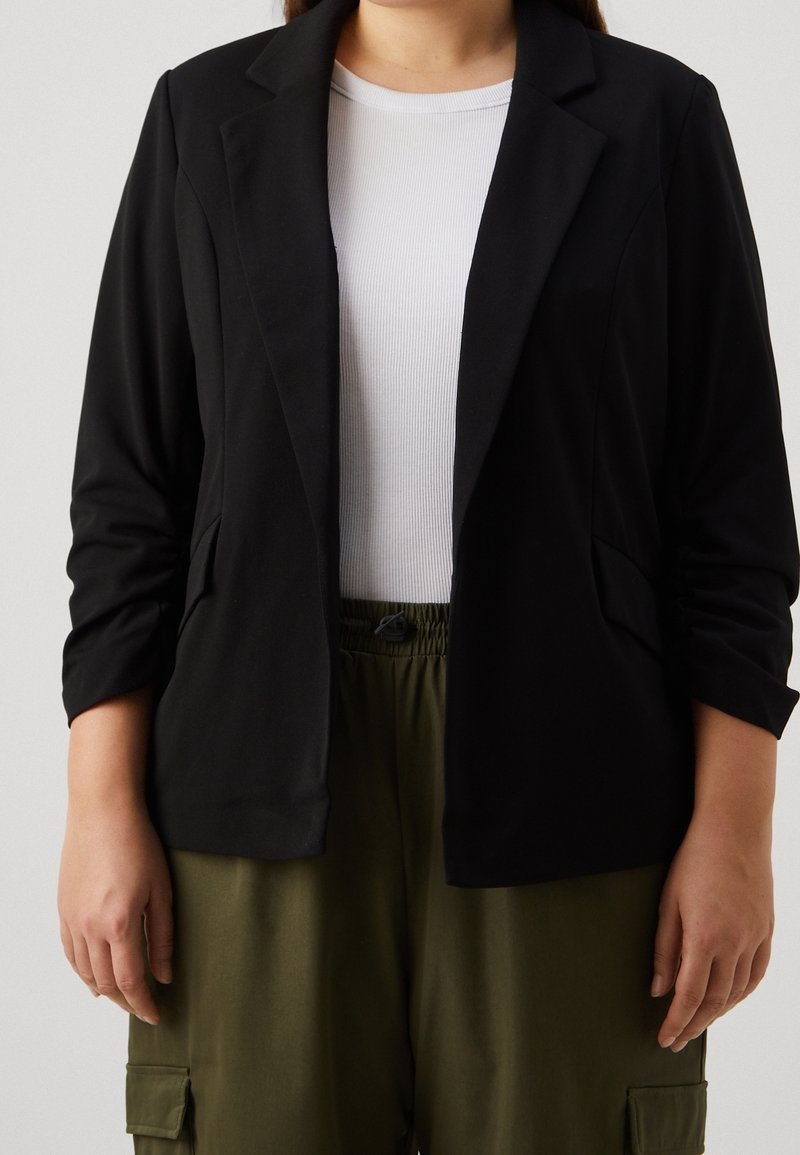 Femme portant un blazer noir sur une chemise blanche côtelée et un pantalon cargo vert olive, debout sur un fond uni.