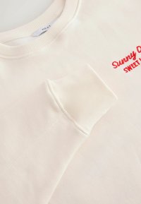 Manche et encolure de sweat-shirt crème avec texte rouge brodé "Sunny D" et "SWEET" sur la poitrine, montrant le détail du poignet côtelé.