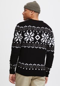 Black knitted sweater featuring white snowflake patterns across the chest and sleeves. Ribbed cuffs and hem. Paired with an olive beanie.