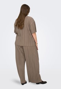 Γυναίκα με μακριά μαλλιά που φοράει χαλαρό, πλισέ τοπ σε αποχρώσεις του ταουπέ και φαρδιά παντελόνια, με την πλάτη γυρισμένη, σε απλό ανοιχτό φόντο.