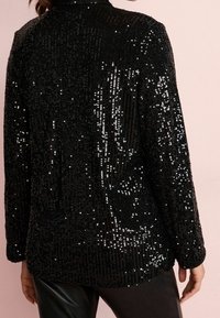 Chaqueta negra de lentejuelas con un patrón tejido texturizado, que presenta un cuello alto y mangas largas. Las lentejuelas crean una superficie reflectante.