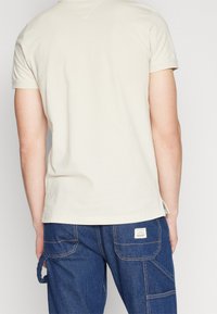 Mann in beige Hemd mit kurzen Ärmeln und blauen Denim-Cargohosen, von hinten gezeigt vor einem schlichten hellen Hintergrund.