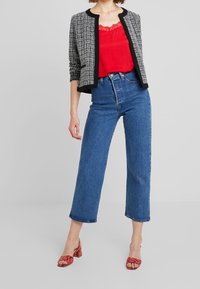 Czarna i biała kurtka z tweedu o okrągłym dekolcie, nałożona na czerwony top na ramiączkach z koronkową lamówką, zestawiona z jeansami z wysokim stanem i czerwonymi sandałami na słupku.