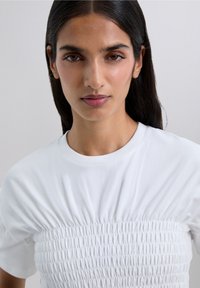 White t-shirt featuring a gathered design at the middle, short sleeves, and a classic crew neckline. Smooth texture and fitted style.